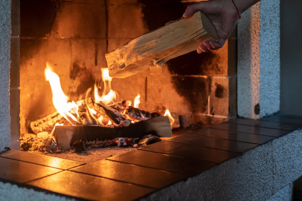 Alimenter son feu de cheminée avec du bois sec Main posant une bûche de bois dans une cheminée en briques. Un feu ardent avec des flammes oranges illumine l'âtre.
