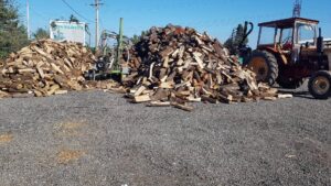 Gros stock de bois de chauffage "BONNADIEU VENTE AU DÉTAIL ÉLAGAGE" avec tracteur rouge et fendeuse sur gravier.