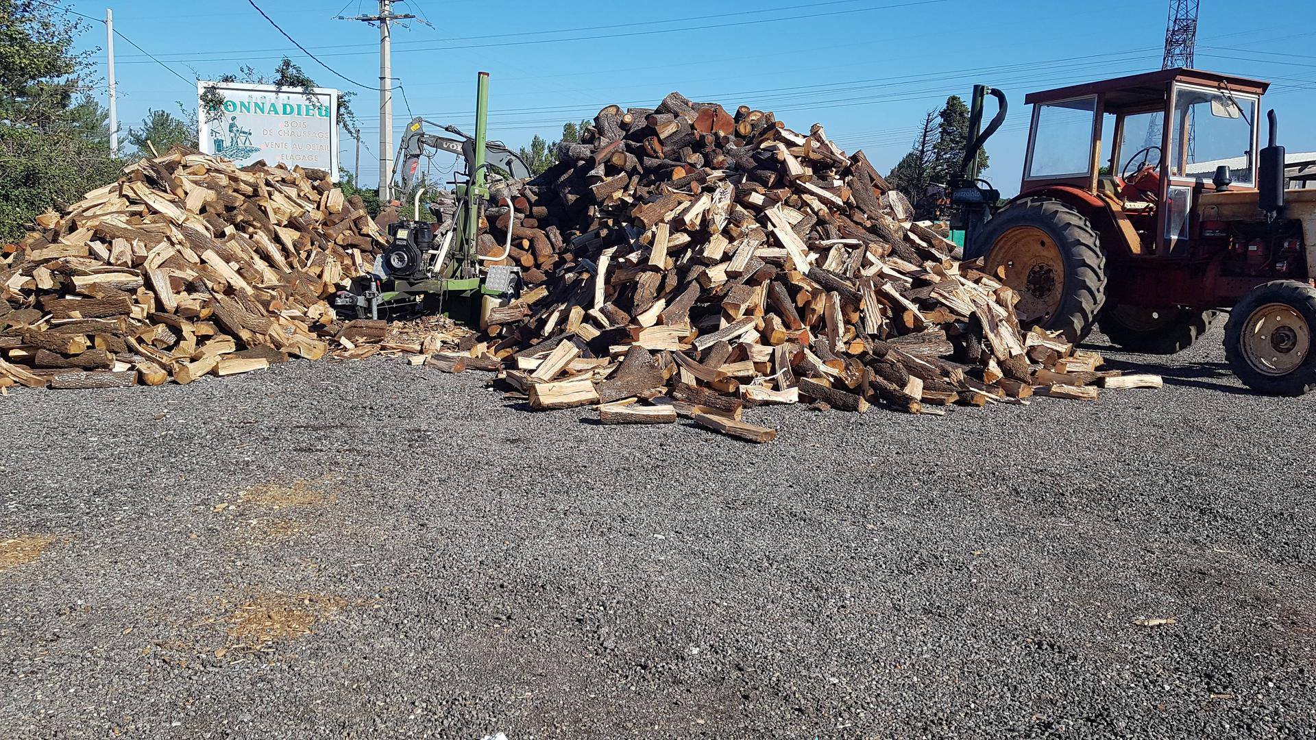 Gros stock de bois de chauffage "BONNADIEU VENTE AU DÉTAIL ÉLAGAGE" avec tracteur rouge et fendeuse sur gravier.