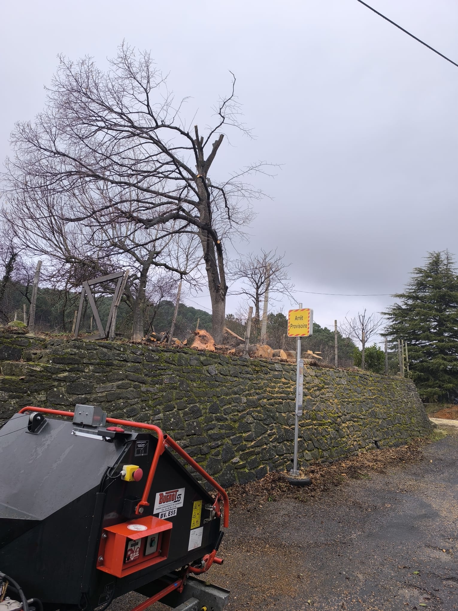 Broyeur Bugnot BV E55 devant un mur de pierre et un panneau Arrêt Provisoire pendant des travaux d'élagage.
