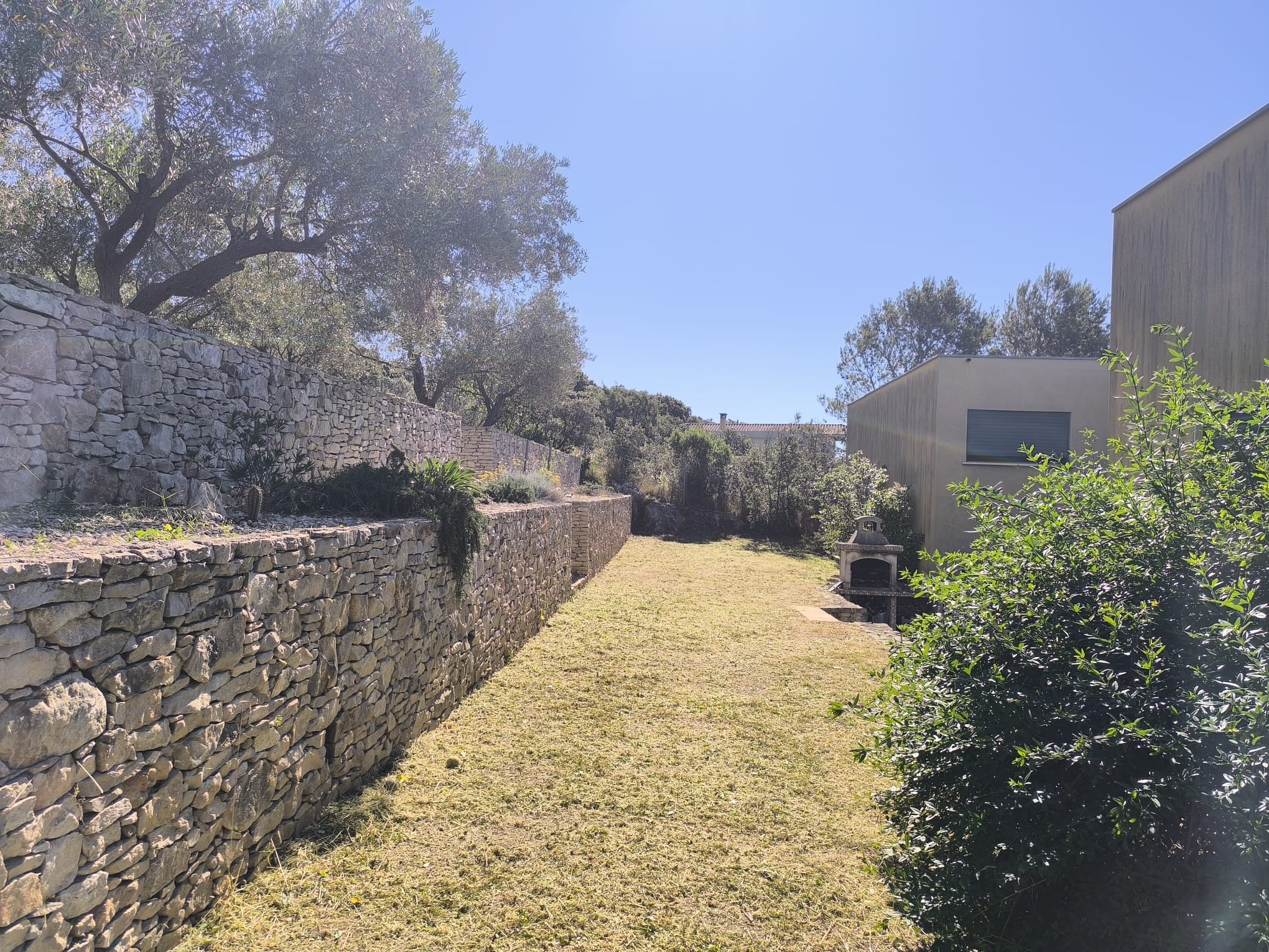 Mur en pierre sèche et jardin ensoleillé avec barbecue extérieur près d'une maison contemporaine.
